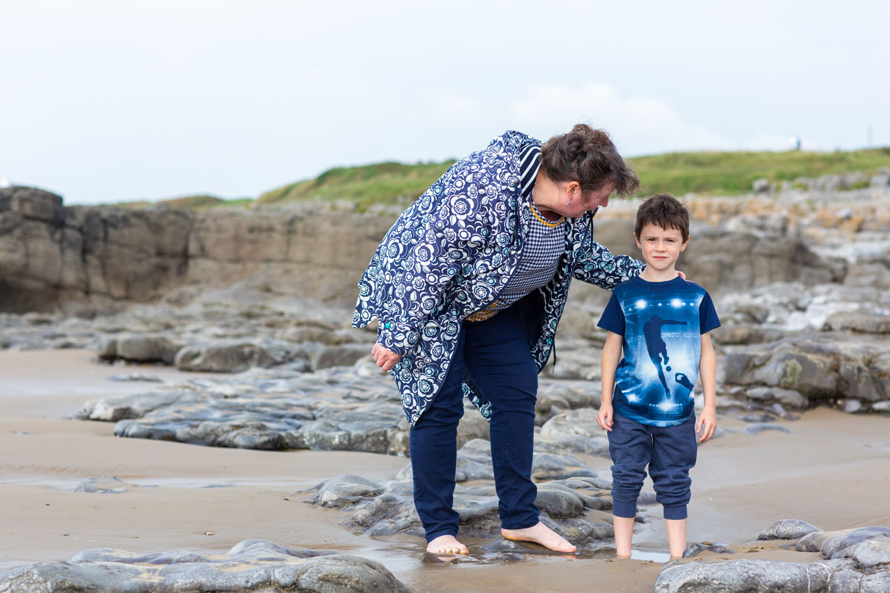 Boy on the beach with trouser legs rolled up. Lady has a reassuring hand on his shoulder.