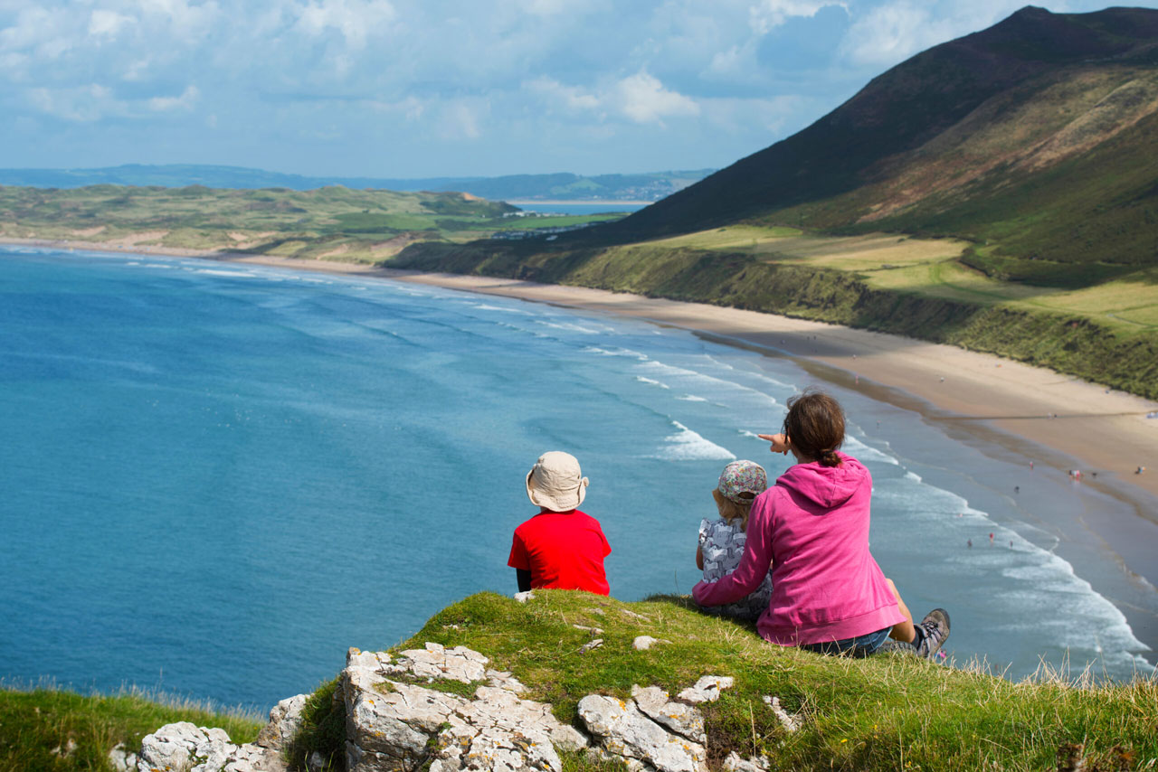 Adult with two young children sitting at top of cliffs looking over coastal bay