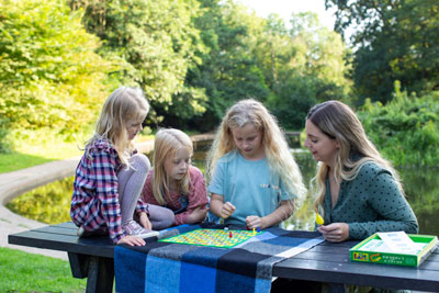Four people play a board game on a picnic table covered with a blue blanket, by a tranquil pond amidst lush greenery.