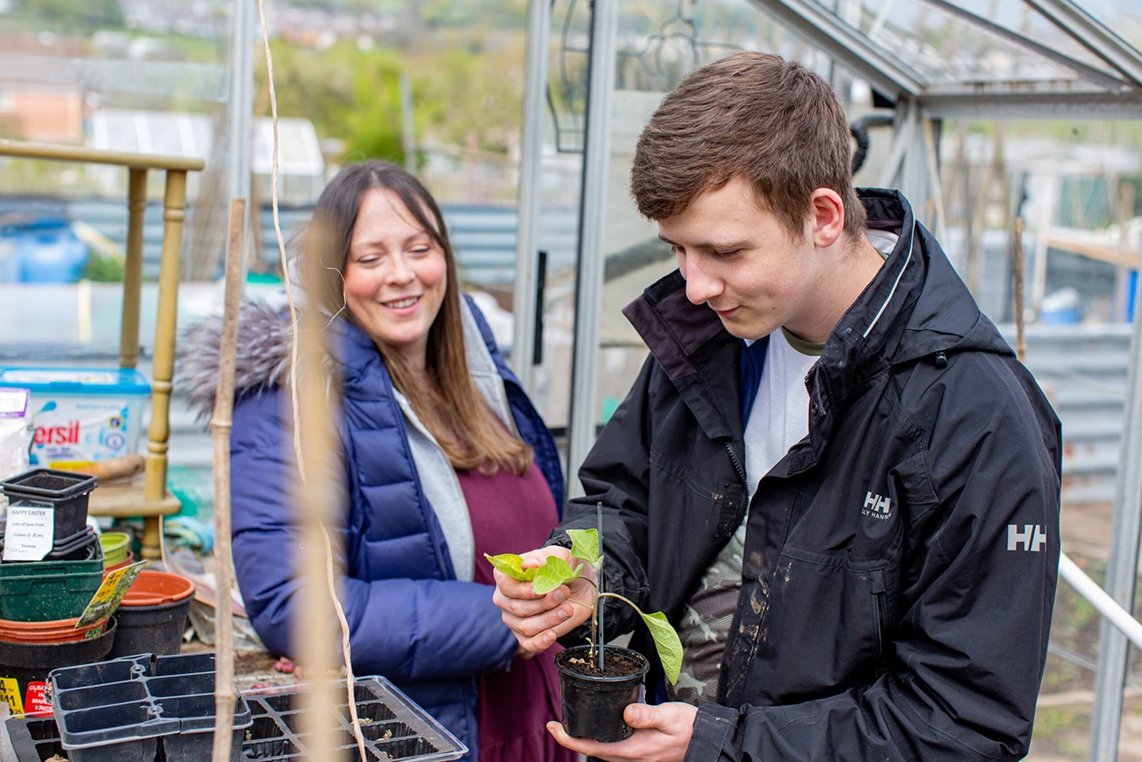 A young man holds a small potted plant, smiling while a woman watches nearby inside a greenhouse. Various gardening supplies and pots surround them, suggesting a shared gardening activity.